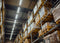 The interior of a warehouse with overhead lights and wood palettes stacked high on metal shelves filled with plastic wrapped brown cardboard boxes.