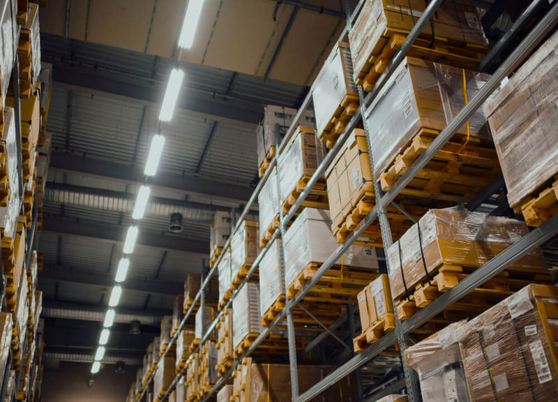 The interior of a warehouse with overhead lights and wood palettes stacked high on metal shelves filled with plastic wrapped brown cardboard boxes.