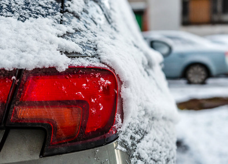 The right rear bumper and light of a grey 4-door Sedan covered with snow and ice on a snow covered street.
